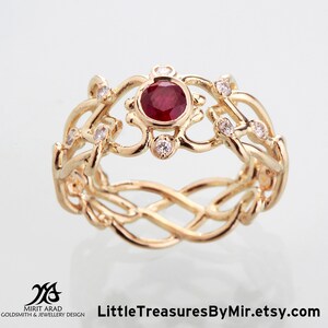 Puede incluir: Un anillo de oro con una piedra preciosa de rubí rojo rodeada de diamantes más pequeños. El anillo tiene un diseño delicado e intrincado con una banda trenzada.