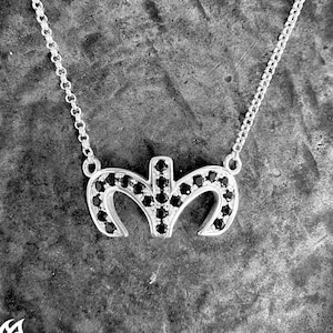 Op de afbeelding: Een zilveren ketting met een hanger in de vorm van de letter 'M' bezet met zwarte edelstenen. De hanger is afgebeeld tegen een getextureerde grijze achtergrond.