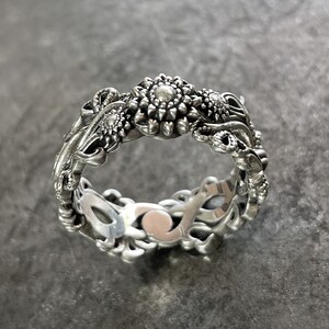May include: A silver ring with a floral design. The ring is made of intricate details and features a central flower with a pearl-like center.
