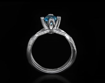 Könnte beinhalten: Ein silberner Verlobungsring mit einem blauen Edelstein, der in einem blumenförmigen Design gefasst ist. Der Ring hat eine strukturierte Band und ist vor einem schwarzen Hintergrund platziert.
