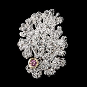 May include: A silver-toned, organic-shaped pendant with a textured surface and a small, round, pink gemstone set in a gold-coloured bezel. The pendant is set against a black background. The bottom left corner has the text "MIRIT ARAD GOLDSMITH & JEWELLERY DESIGN".