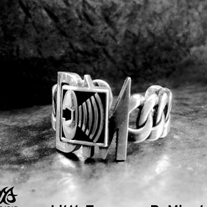 Puede incluir: Un anillo de plata con un símbolo de altavoz negro y plateado y un diseño de rayo plateado. El anillo está sobre una superficie gris texturizada. El texto "LittleTreasuresByMir.etsy.com" es visible en la imagen.