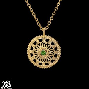 Pode incluir: Um colar de ouro com um pingente redondo que apresenta um design delicado e intrincado e uma pequena gema verde no centro.