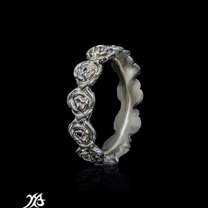 Puede incluir: Un anillo de plata con una banda de pequeñas rosas alrededor de la parte superior. La banda es texturizada y tiene un borde festoneado.