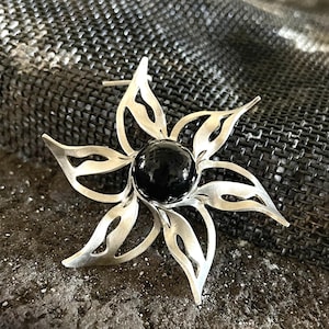 Puede incluir: Broche plateado con forma de flor y una piedra negra en el centro. El broche tiene seis pétalos calados y un cierre de alfiler. La joya se exhibe sobre una superficie texturizada de color gris oscuro.
