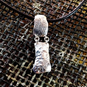 May include: A silver necklace with a textured pendant hangs on a black leather cord. The pendant is made of two pieces, one on top of the other, connected by small silver rings. The necklace is displayed on a textured metal surface.