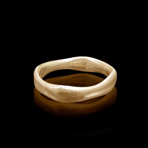 Puede incluir: Un anillo de banda de oro simple con una forma ligeramente irregular y orgánica. El anillo es liso y pulido, con un brillo sutil.