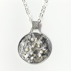 Puede incluir: Un collar con colgante de plata que presenta un dije circular con una superficie texturizada y oscura oxidada. El colgante está adornado con pequeñas perlas esféricas blancas y diminutos detalles en tono dorado. Cuelga de una cadena de plata.
