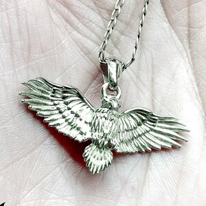 Puede incluir: Collar con colgante de águila de plata con un diseño detallado. El águila está en vuelo con las alas extendidas. La cadena es una cadena de plata delicada.