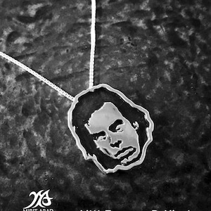 Puede incluir: Un collar de plata con un colgante que presenta una silueta en blanco y negro de la cara de una persona. El colgante tiene forma de cabeza y tiene un contorno detallado de los rasgos de la persona. El collar está sobre un fondo oscuro texturizado.
