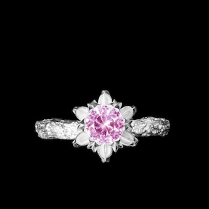 Puede incluir: Un anillo de plata con una piedra preciosa rosa engastada en un diseño con forma de flor. La banda del anillo es texturizada y tiene un aspecto natural y orgánico.