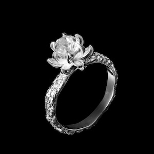 Puede incluir: Un anillo de plata con un engaste en forma de flor y una piedra preciosa transparente. La banda es texturizada y tiene un aspecto rústico.