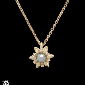 Pode incluir: Um colar de ouro com uma corrente delicada e um pingente em forma de flor com uma pérola azul claro no centro.
