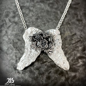 May include: A silver necklace with a pendant shaped like two wings with a cluster of black flowers in the centre.