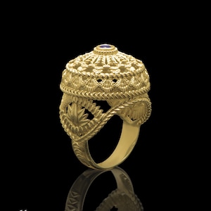 Puede incluir: Un anillo de oro con un diseño detallado e intrincado. El anillo presenta una parte superior en forma de cúpula con una pequeña piedra preciosa azul en el centro.