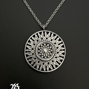 Puede incluir: Un collar con un colgante de plata de diseño circular, que presenta un patrón de rayos de sol y un diseño floral más pequeño en el centro.
