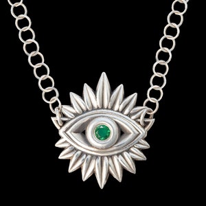 May include: Silver necklace featuring a green gemstone eye pendant. The eye is surrounded by a sunburst design.