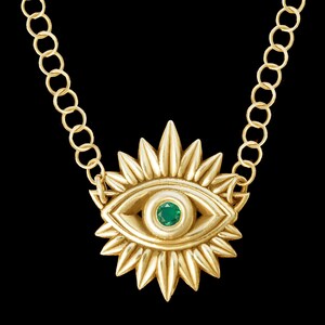 Puede incluir: Un collar de oro con un colgante en forma de ojo con una piedra preciosa verde en el centro. El ojo está rodeado por un diseño de rayos de sol.