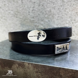 May include: A black leather wrap bracelet with two silver charms. One charm is oval shaped with a delicate plant design. The other charm is rectangular with a textured surface.