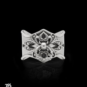 Könnte beinhalten: Ein silberner Ring mit einem verzierten Design, das ein stilisiertes Blumenmotiv und ein ringförmiges Detail aufweist. Der Ring ist aus Sterlingsilber gefertigt und hat eine polierte Oberfläche.