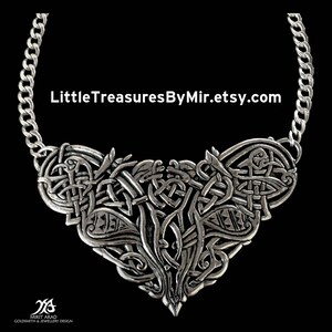 Op de afbeelding: Een zilveren ketting met een hartvormige hanger in Keltische knoopvorm. De hanger is ingewikkeld ontworpen met een knoopwerkpatroon en heeft aan elke kant een kleine vogel. De ketting is gemaakt van een zilveren ketting.