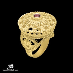 Puede incluir: Un anillo de oro con un diseño detallado e intrincado que presenta una piedra preciosa roja en el centro. El anillo está hecho de oro de 18 quilates y es una pieza llamativa.