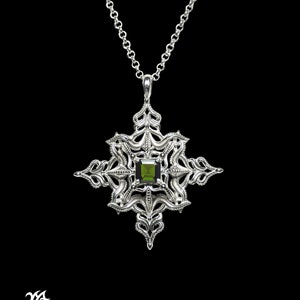 May include: A silver pendant necklace featuring a green gemstone at its centre. The pendant is shaped like a star with elaborate detailing. The necklace is displayed against a black backdrop.