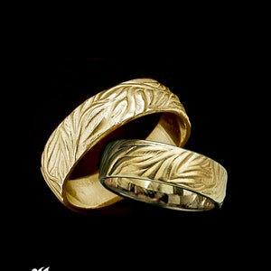 May include: Two gold wedding bands with a leaf pattern. The bands are stacked on top of each other.