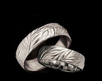 Puede incluir: Dos alianzas de plata con un diseño de hojas. Las alianzas están entrelazadas y la imagen está sobre un fondo negro.