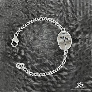 Puede incluir: Un brazalete de cadena de plata con un delicado dije ovalado que presenta un diseño de rosa recortada. El dije está unido a la cadena con un pequeño lazo.