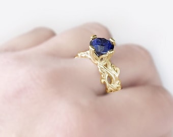 May include: A gold ring with a blue gemstone set in a delicate, intricate design. The ring is on a hand, showcasing the detail of the band.