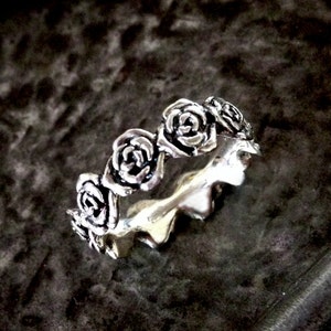 Könnte beinhalten: Ein silberner Ring mit einem sich wiederholenden Rosenmuster. Der Ring zeigt ein detailliertes Blumendesign mit mehreren kleinen Rosen. Er wird auf einer dunklen, strukturierten Oberfläche mit unscharfem Hintergrund präsentiert.
