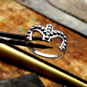 May include: A silver ring with a textured design in the shape of a crown. The ring is being held by tweezers.