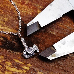 Könnte beinhalten: Eine silberne Halskette mit einem Anhänger in Form des Buchstabens "J" mit kleinen Diamanten. Die Halskette wird mit einer silbernen Schieblehre gemessen.