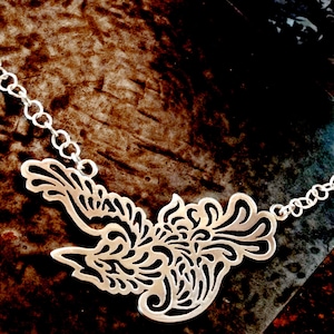 Op de afbeelding: Een zilveren ketting met een gedetailleerde, ingewikkelde vogelhanger. De hanger is gemaakt van zilver en heeft een delicaat, filigraan ontwerp. De ketting ligt op een donkerbruin oppervlak met een tang en een mok met een zwart-wit afbeelding van een persoon.