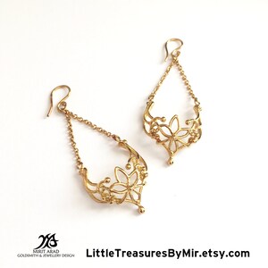 K&ouml;nnte beinhalten: Goldene Ohrh&auml;nger mit einem zarten floralen Design. Die Ohrringe haben eine Kette mit einem blumenf&ouml;rmigen Anh&auml;nger.