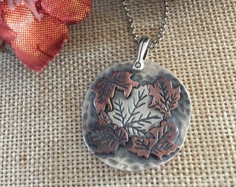 May include: A round, hammered silver-tone pendant necklace featuring a central silver maple leaf surrounded by four smaller copper-colored leaves. The pendant hangs from a silver-tone ball chain, showcasing a mixed-metal design with textured details.