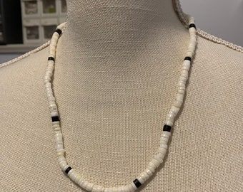 Op de afbeelding: Een witte kralenketting met zwarte accentkralen. De ketting wordt op een mannequin getoond. De kralen zijn klein en rond, geregen om een eenvoudig, elegant ontwerp te vormen. De ketting heeft een neutrale kleur en kan bij veel outfits worden gedragen.