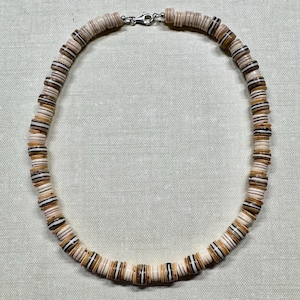 May include: A necklace made of small, flat, shell-like beads in shades of cream, brown, and black. The beads are strung together, forming a circular shape. The necklace has a silver-colored clasp.