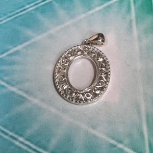 May include: A silver-toned oval pendant featuring an intricate openwork filigree pattern throughout the frame. The outer edge is defined by a delicate beaded texture, and a polished bail is attached to the top for hanging on a chain or cord.