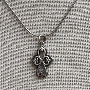 Peut inclure: Un collier argenté composé d'une fine chaîne serpent et d'un pendentif en forme de croix avec des motifs de volutes ajourés. Le métal présente une finition polie avec des zones en creux assombries qui soulignent les détails ornementaux.