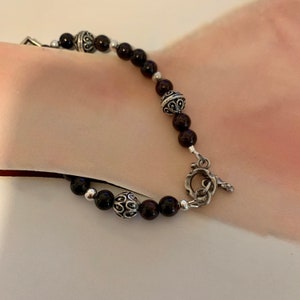 May include: A beaded bracelet featuring dark red, round beads and ornate silver-toned metal accents. The bracelet has a toggle clasp and is displayed on a light background. The bracelet is a piece of jewelry.