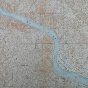 May include: A vintage topographic map featuring Lake Chelan in Washington State. The map shows contour lines, elevation details, and the surrounding mountainous terrain. The lake is a light blue color, and the land is a mix of tan and brown.