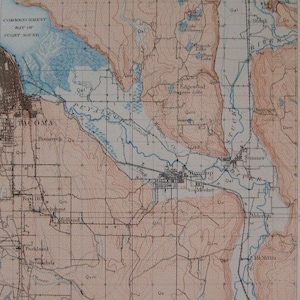 May include: A vintage map of Tacoma, Washington, showing the Commencement Bay of Puget Sound, the Puyallup River, and surrounding towns. The map is printed in black and white with blue lines for water features and red lines for elevation.
