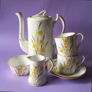 Könnte beinhalten: Ein weißes Porzellan-Tee-Set mit einem Blumendesign mit gelben Narzissen. Das Set enthält eine Teekanne, eine Milchkännchen, eine Zuckerdose und zwei Teetassen mit Untertassen.