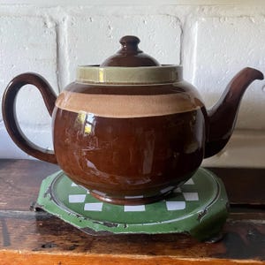 Può includere: Una teiera in ceramica marrone lucido con corpo arrotondato, manico curvo e coperchio abbinato. La sezione superiore presenta un'ampia fascia orizzontale in una tonalità marrone chiaro, sormontata da un bordo color verde oliva spento.