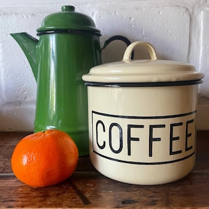 May include: A green enamel coffee pot and a cream-colored coffee canister with black trim and the word "COFFEE" in black block letters. An orange tangerine sits in front of the items on a wooden surface.