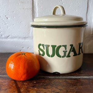 May include: Cream enamel sugar storage jar with a lid and green "SUGAR" lettering. A bright orange tangerine sits beside the jar on a wooden surface. The jar has a black rim and a small handle on the lid.