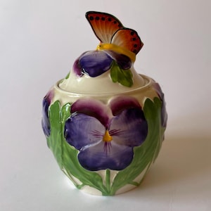May include: A decorative ceramic jar with a lid, adorned with a butterfly and pansy flower motif. The butterfly is orange and brown, resting on a purple pansy. The jar is cream-coloured, with purple pansies and green leaves painted around the sides.