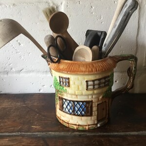 Puede incluir: Una jarra de cerámica con forma de cabaña con un techo marrón y un asa. La jarra está llena de cucharas de madera, un cepillo y un par de tijeras.